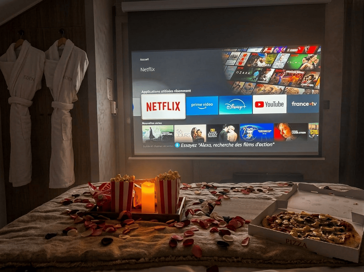 Écran géant avec Netflix, peignoirs Eden, popcorn et pizza sur le lit